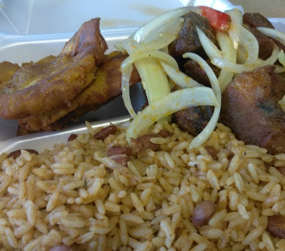 Griot with onions, rice and beans, and fried plantain in a takeout box